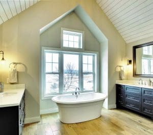 Spacious bathroom with standalone bathtub, wood flooring, and four large energy-efficient windows overlooking landscape.