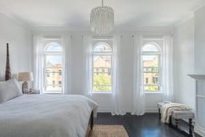 Spacious bedroom with wood flooring, white walls, and three specialty windows outfitted with white window treatments.