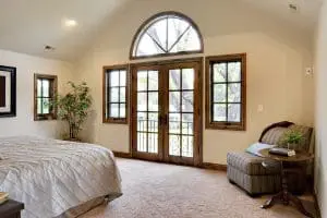 Interior view of home with carpeting, large bed, chair in corner, and wood-frame hinged patio doors leading to the outdoors.