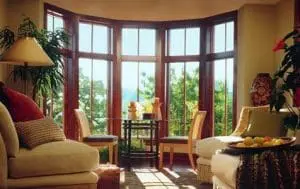 View of interior of home on a sunny day. Room features four chairs, one side table, and a tall bow window made up of five narrow windows.