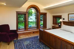 Cozy bedroom with wood floors, wood-framed bed, and custom wood-framed windows that overlook a backyard.