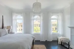 Spacious bedroom with wood flooring, white walls, and three specialty windows outfitted with white window treatments.